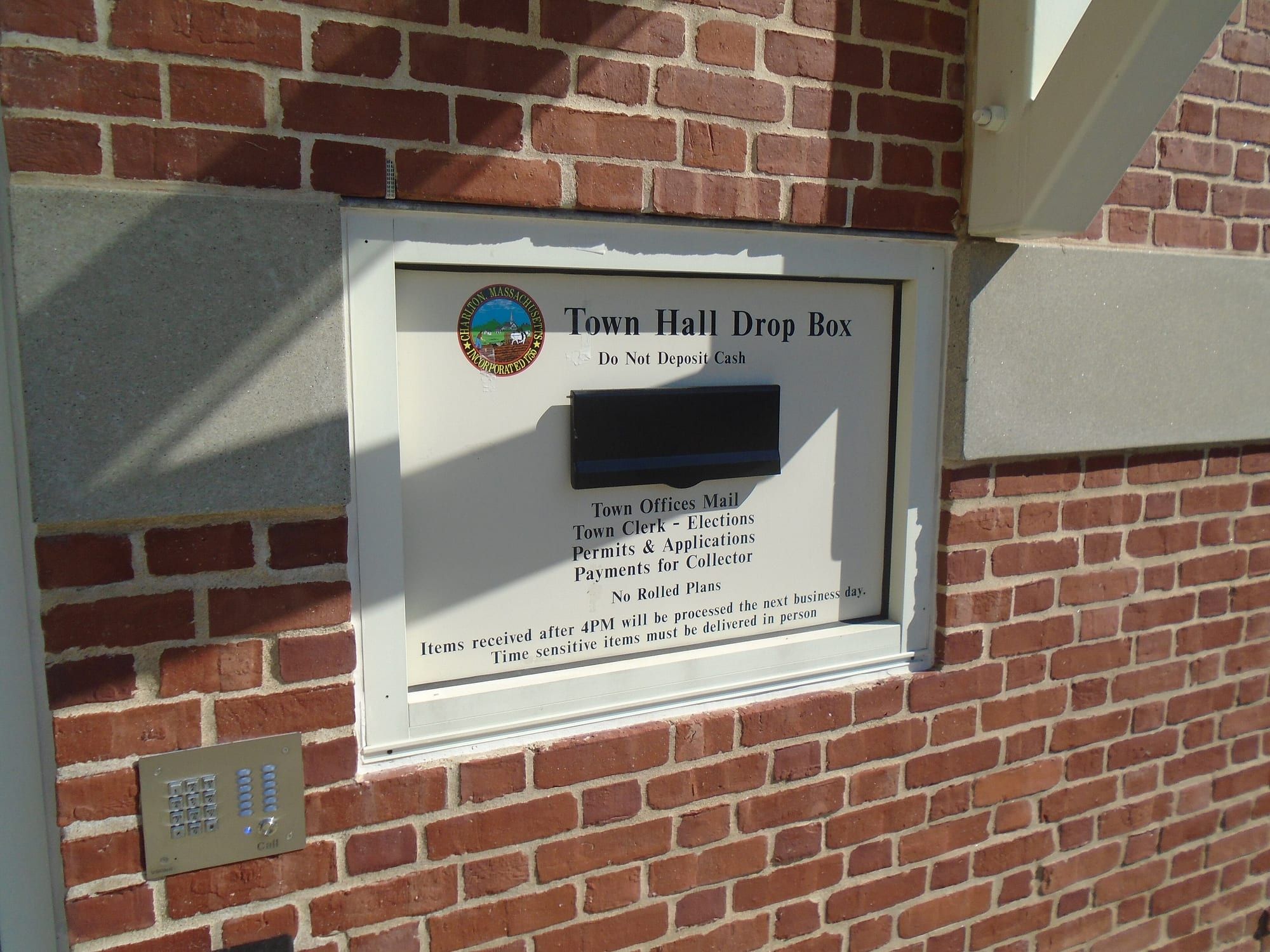A metal drop box set into a brick wall beside a door, with a slot for depositing mail and ballots.