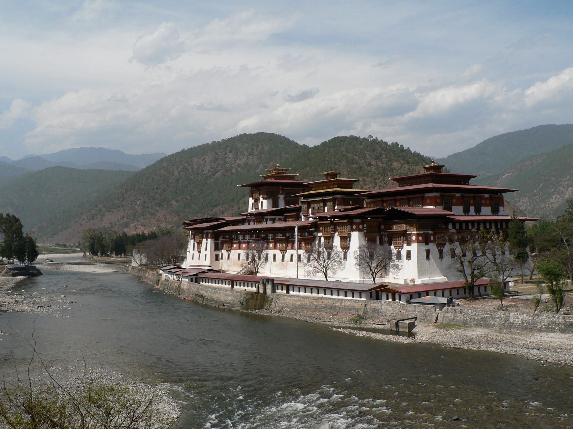 Punakha Dzong, a large white fortress-monastery with red-trimmed roofs, beside a river in a green Himalayan valley under a cloudy sky.