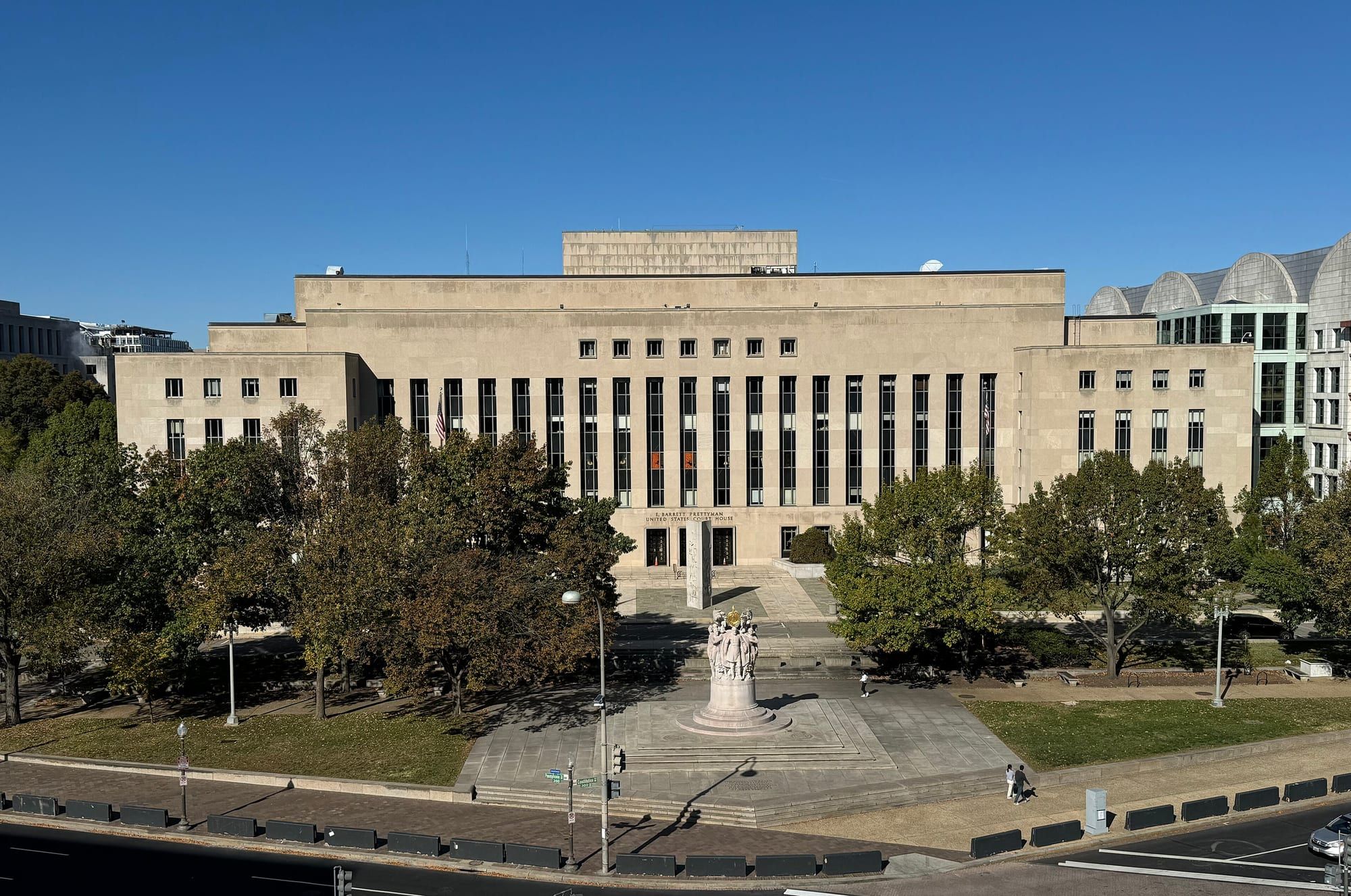 A wide, light-colored stone federal courthouse with rows of tall narrow windows, seen from above across a plaza with trees under a blue sky.