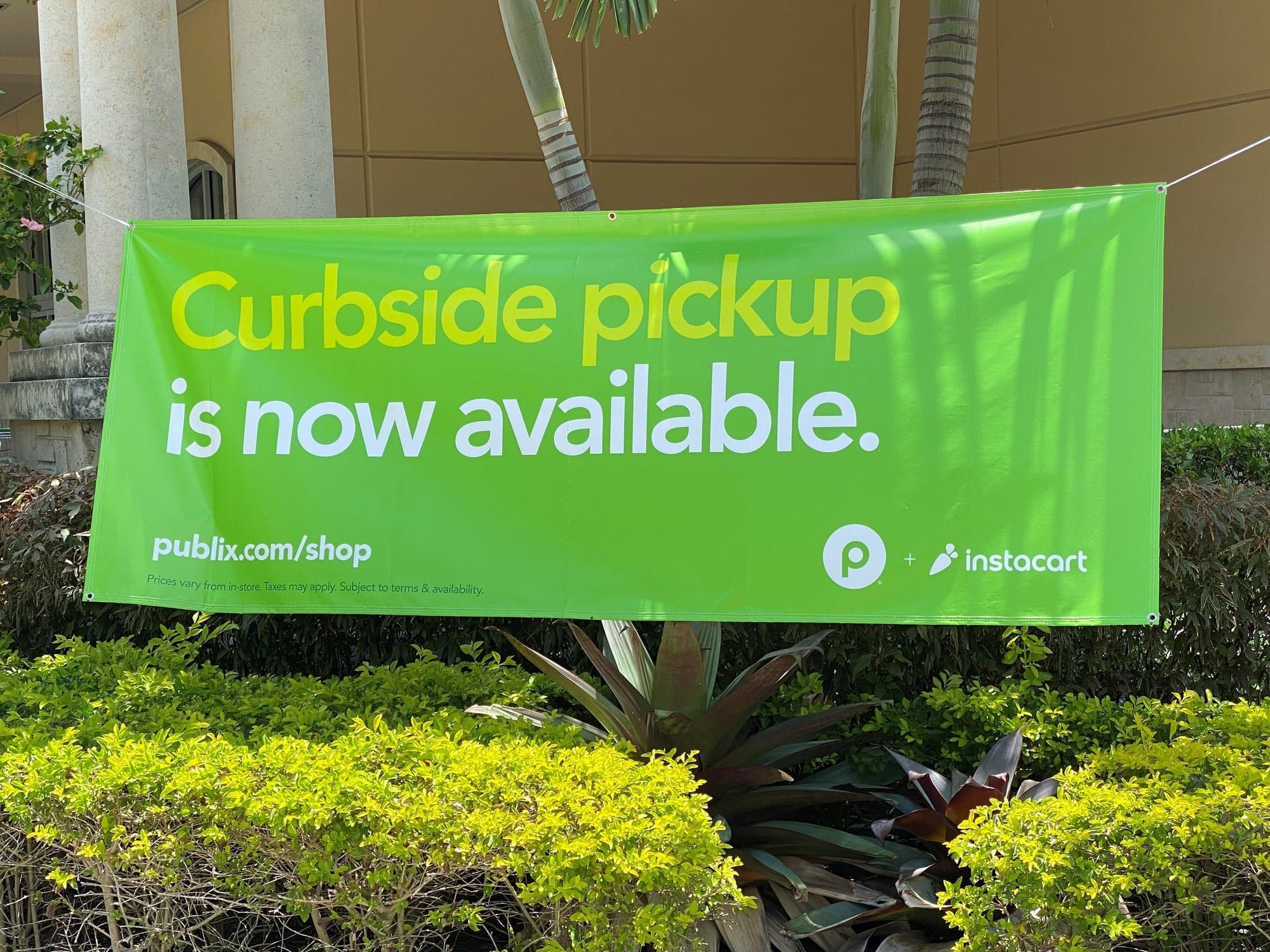 A bright green banner hung under a building's overhang reading “Curbside pickup is now available,” with a supermarket website address and the Instacart logo, above green shrubs.
