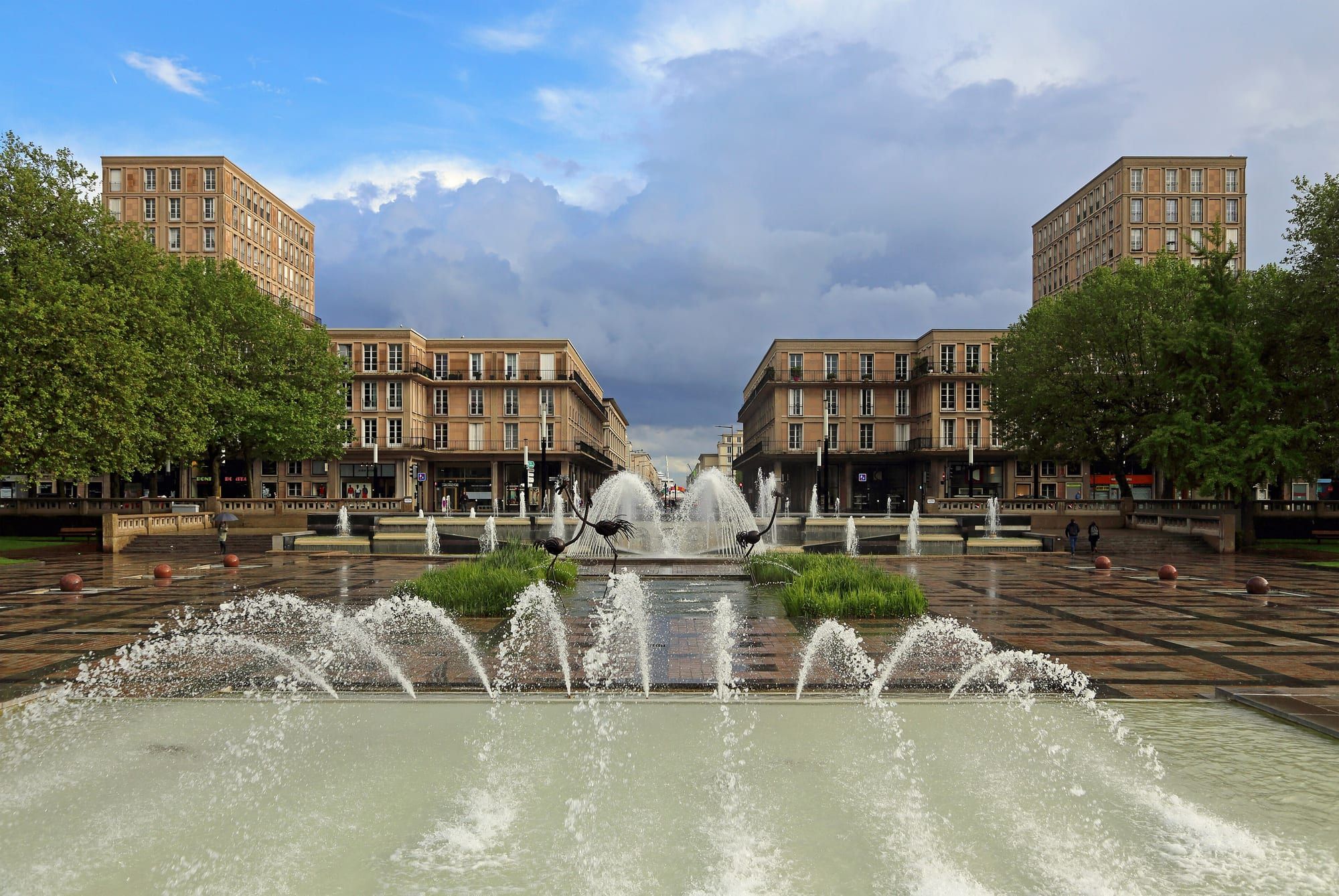 Une grande place pavée avec des jets d'eau et un bassin, bordée d'arbres et d'immeubles en béton clair de l'après-guerre, sous un ciel nuageux.