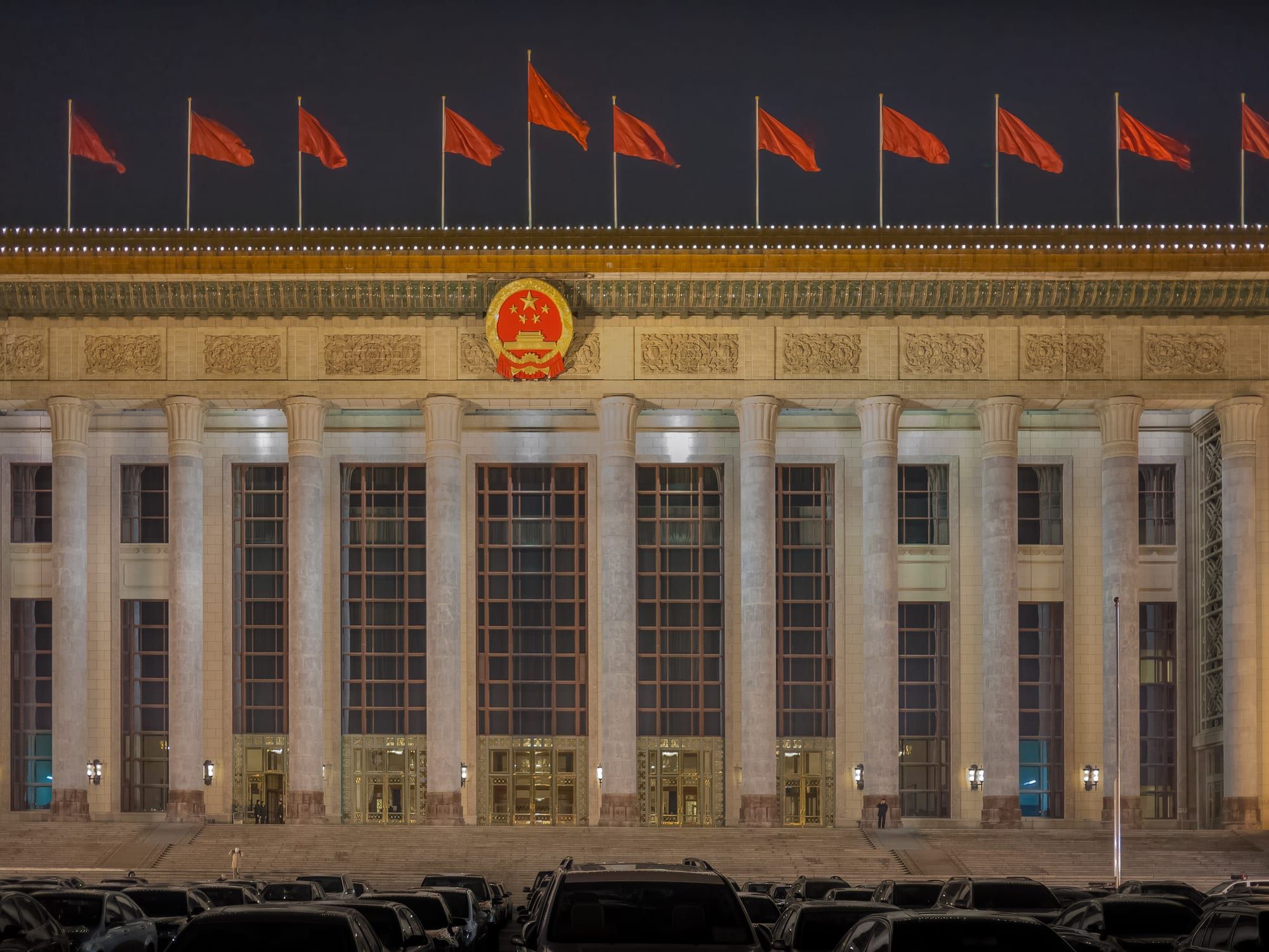 夜间灯光下的北京人民大会堂正立面，楼顶有一排红旗，中央悬挂国徽。