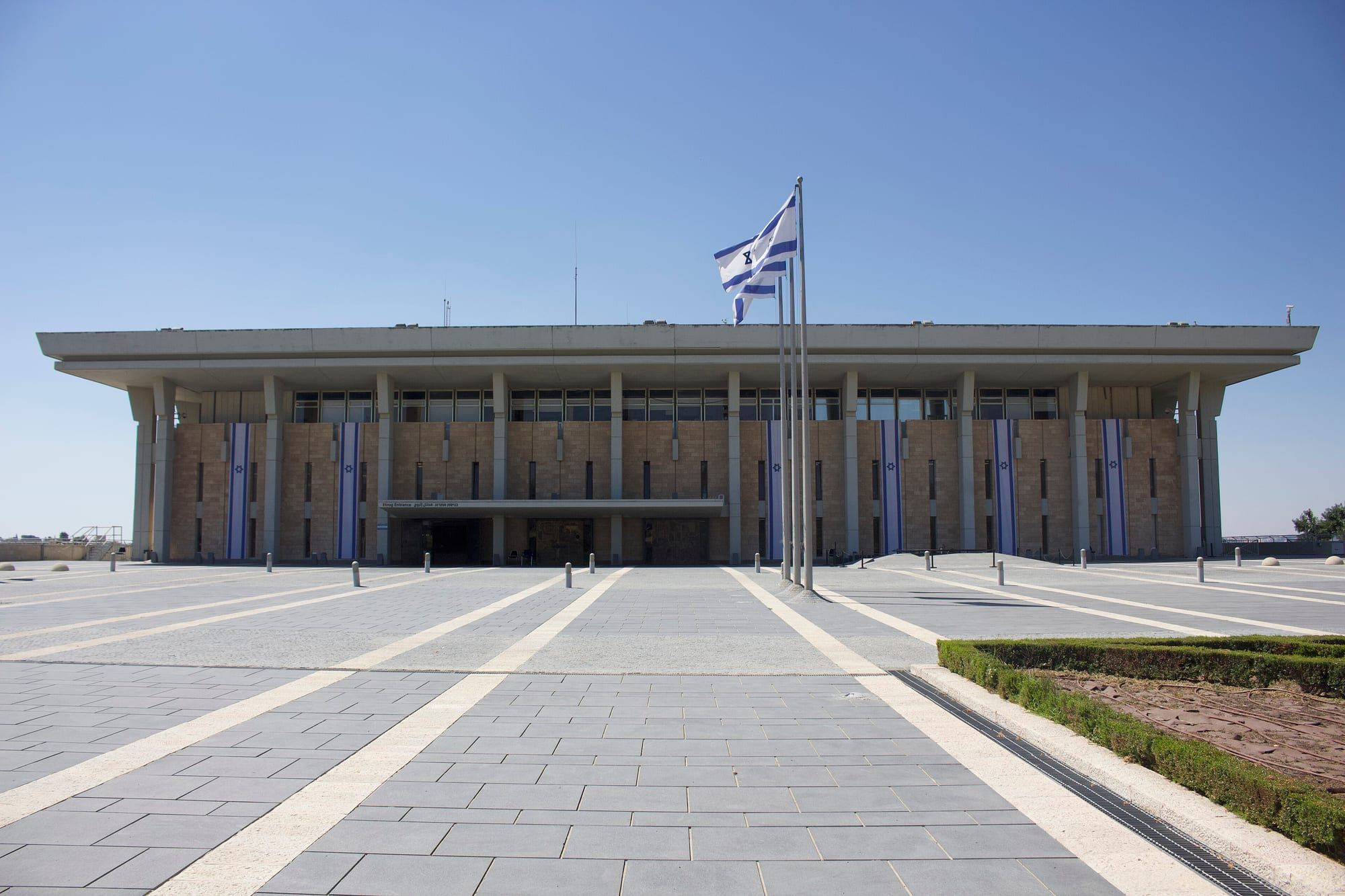 The wide, low concrete and stone façade of the Knesset building, with an Israeli flag on a pole above it and a paved plaza in front.