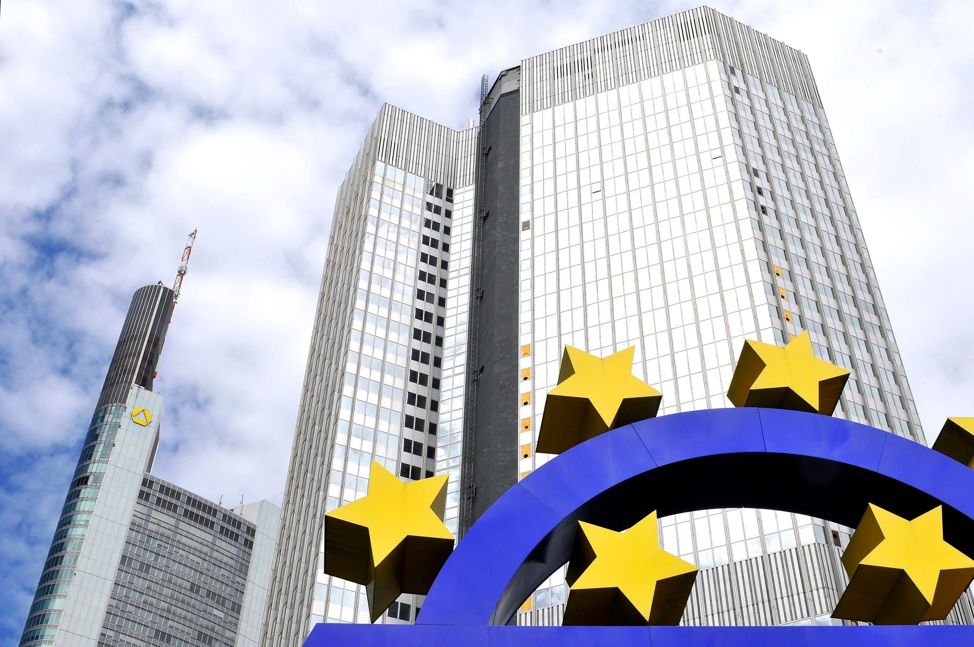 A large blue euro sign sculpture ringed with yellow stars in front of tall glass office towers under a partly cloudy sky.