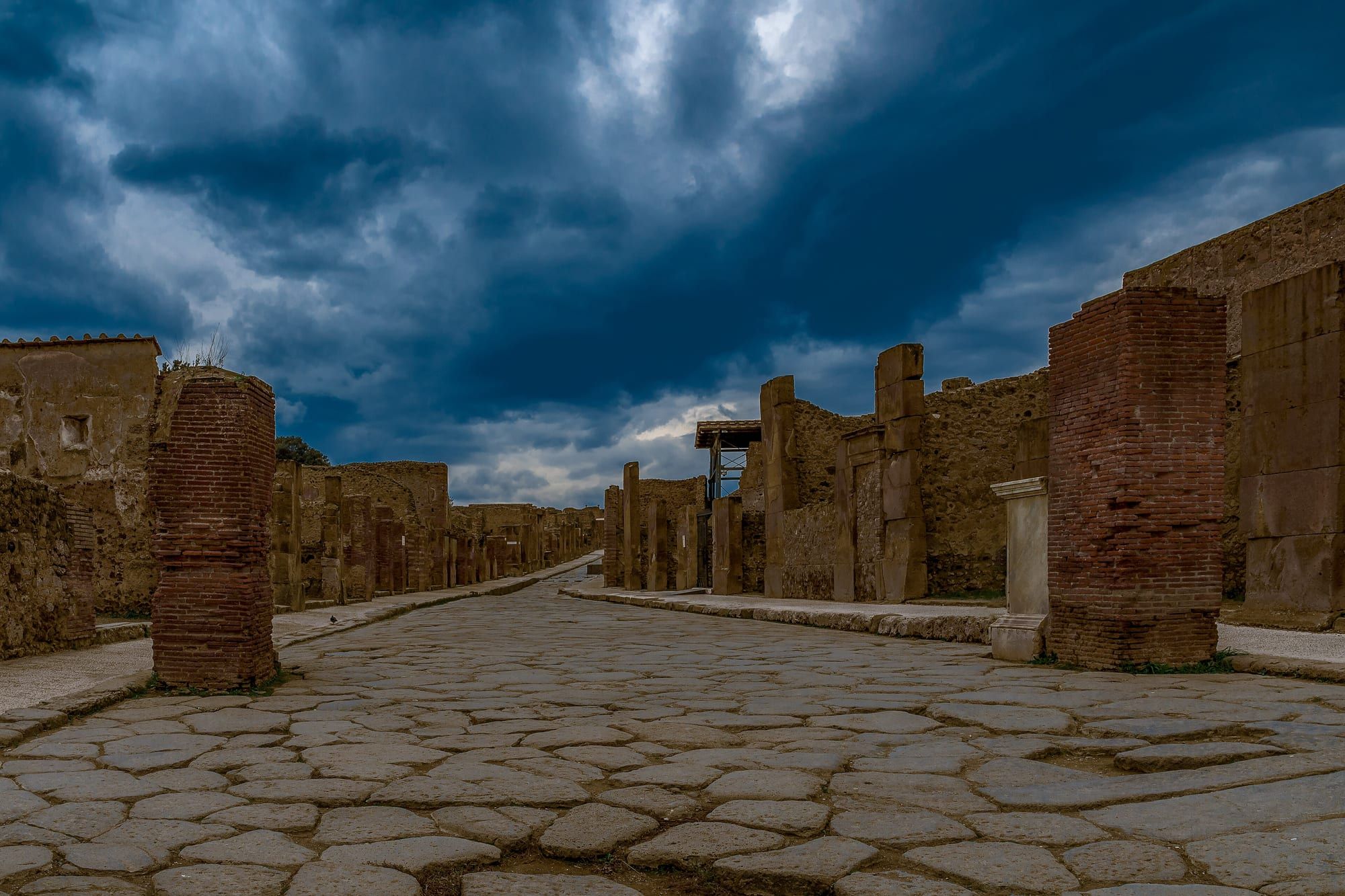 Une rue antique pavée de grandes dalles, bordée de murs de briques et de pierres en ruine, sous un ciel d'orage.