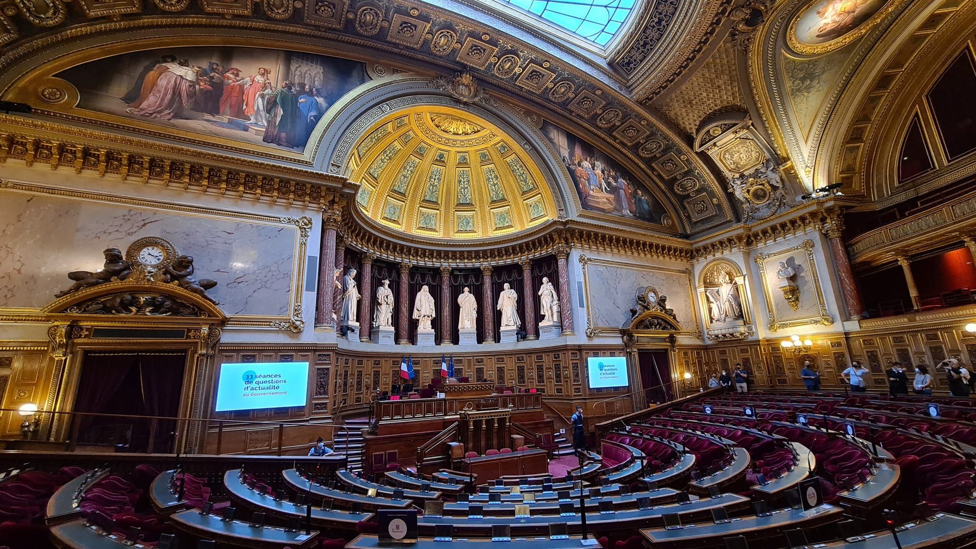 L'hémicycle du Sénat, au palais du Luxembourg : rangées de sièges rouges en demi-cercle, boiseries, statues et coupole dorée.