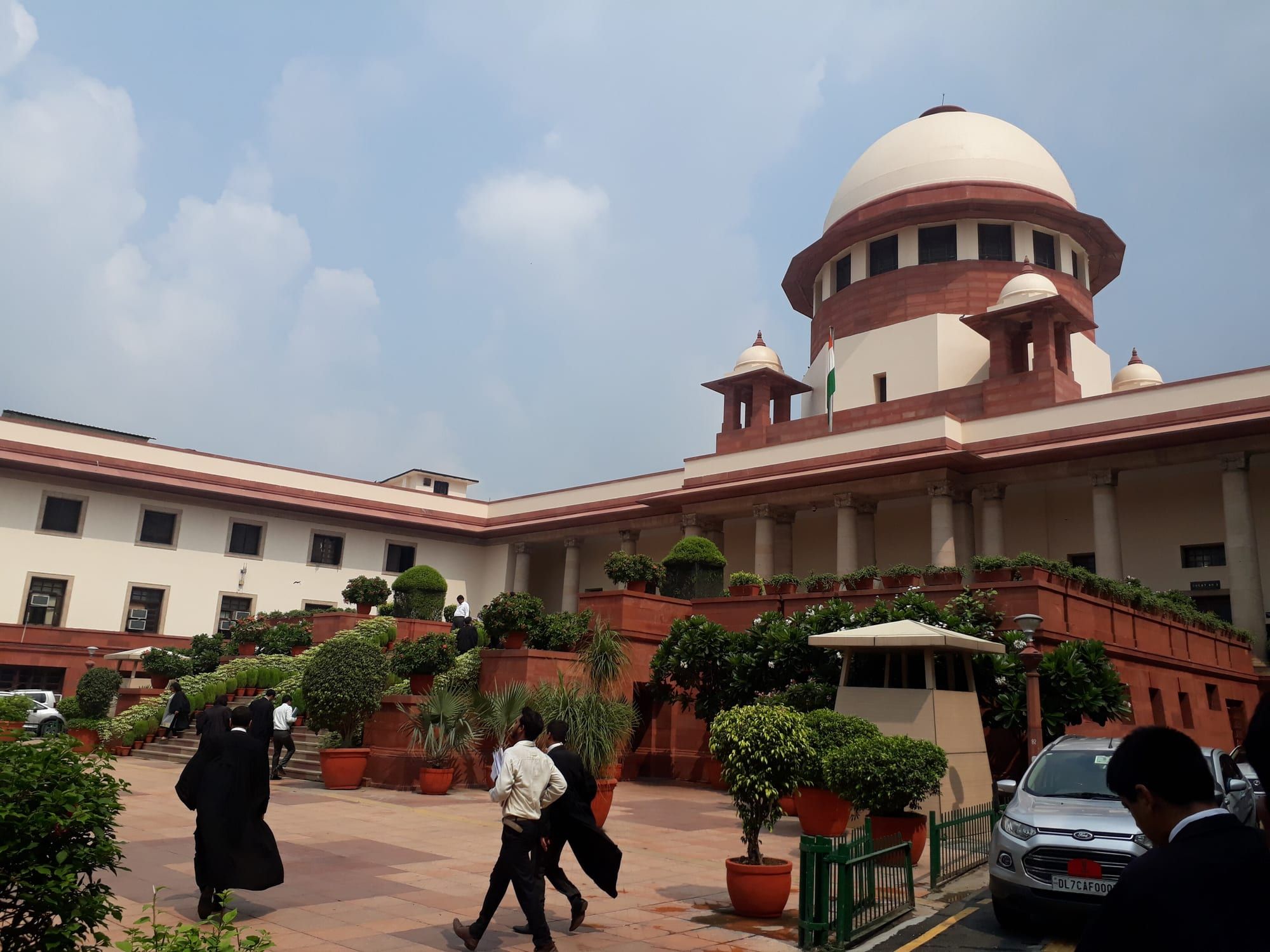 The white dome and red-and-cream colonnaded facade of the Supreme Court of India, seen from its grounds, with trees and parked cars in the foreground.